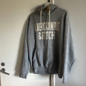 Grå hoodie från Abercrombie & Fitch - Säljer en grå hoodie från Abercrombie & Fitch med broderad logga framtill. Tröjan har  snörning,  och känguruficka Perfekt för alla dagar !