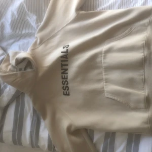 Beige Essentials hoodie. - Snygg beige hoodie från Fear of God Essentials storlek M. Tröjan är använd så det finns ett märke på tröjan så ni ser på bilden, samt börjar bli nopprig på armarna lite 
