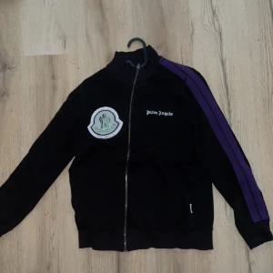 Svart Moncler x Palm Angels ziptröja S - Svart ziptröja från Moncler x Palm Angels med lila ränder längs ärmarna och stor broderad Moncler-patch på bröstet. Palm Angels-logga broderad på andra sidan. Hög krage och hel dragkedja framtill. Perfekt för dig som gillar streetwear med premiumkänsla.