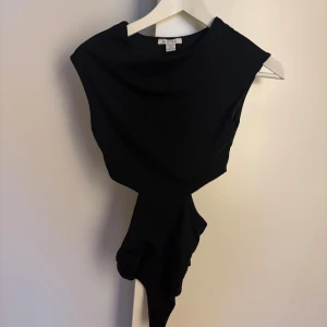 Svart body - Snygg svart body från Amisu med ärmlös design och cool asymmetrisk knytning i midjan. Toppen har en stilren look med hög halsringning och passar perfekt till jeans eller kjol för en trendig outfit.