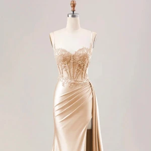 Champagnefärgad långklänning med spets - Säljer en elegant långklänning i champagnefärg med smala axelband och v-ringad byst. Klänningen har ett detaljerat spetsliv med broderade blommor och paljetter, samt en draperad kjol i glansigt satinliknande material och hög slits. Perfekt för bal eller festligheter.