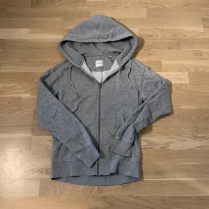 Grå hoodie från Jack & Jones XS - Grå hoodie från Jack & Jones i storlek XS. Tröjan har huva med snörning, dragkedja framtill och ribbade muddar vid ärmar och nederkant. Enkel och stilren design utan mönster.