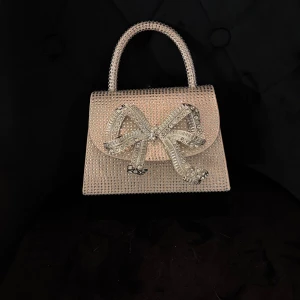 Glittrig handväska med rosett - Superfin glittrig handväska i silver med massor av små stenar och en stor dekorativ rosett framtill. Väskan har ett stadigt handtag och medföljande axelrem i kedja. Perfekt för dig som vill sticka ut och addera extra bling till din look.