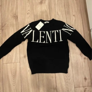 Svart stickad tröja från Valentino - Snygg svart stickad tröja från Valentino med stort vitt logotyp-tryck över bröstet och axlarna. Tröjan har rund halsringning och långa ärmar, perfekt för dig som vill sticka ut med en exklusiv look.