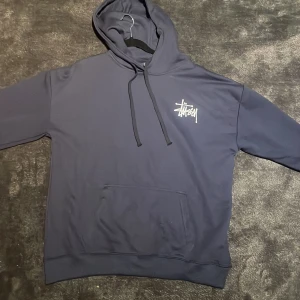 Mörkblå Stüssy hoodie med logga - Säljer en mörkblå hoodie från Stüssy i storlek M. Tröjan har en stor vit Stüssy-logga på ryggen och en mindre logga på bröstet. Klassisk känguruficka framtill och huva. Materialet är mjuk bomull, perfekt för chill dagar.