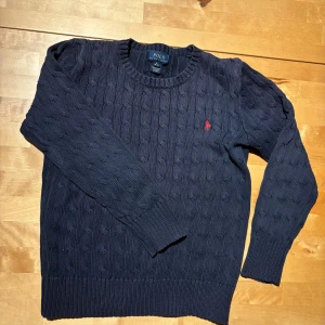Mörkblå stickad tröja Polo Ralph Lauren - Kabelstickad mörkblå tröja från Polo Ralph Lauren med klassisk rund halsringning och röd broderad logga på bröstet. Tröjan har långa ärmar och ribbade muddar för en snygg passform. Perfekt för dig som gillar stilren och tidlös design.