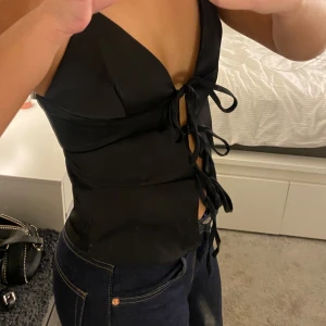 Svart snörad satin topp - Snygg svart topp med smala axelband och djup v-ringning. Toppen har snörning framtill med flera knyt och har ett glansigt satin material. Helt oanvänd!