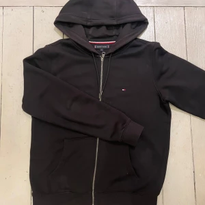 Svart hoodie från Tommy Hilfiger - Svart hoodie från Tommy Hilfiger med dragkedja, huva och klassisk logga på bröstet. Tröjan har fickor framtill och ribbade muddar. Snygga röda och vita detaljer på insidan av luvan. Perfekt för chill dagar eller när du vill ha en clean look.