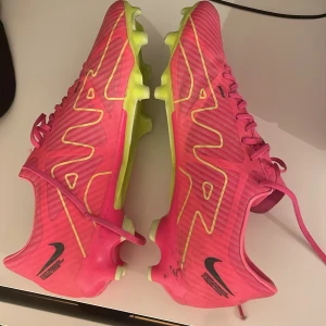 Nike Air Zoom rosa fotbollsskor strl 44 - Säljer ett par Nike Air Zoom fotbollsskor i en riktigt snygg rosa färg med gula detaljer och sula. Skorna har snörning och är designade för gräsplan, med en lätt och smidig syntetöverdel. Perfekta för dig som vill sticka ut på planen.