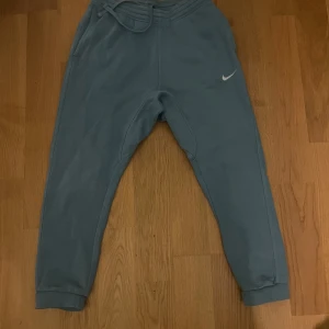 Blå mjukisbyxor från Nike, strl M - Snygga blå mjukisbyxor från Nike med dragsko i midjan och klassisk logga på låret. Byxorna har muddar vid bensluten och en bakficka med knapp. Perfekta för chill eller träning.