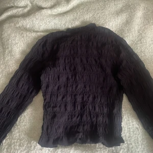 Svart smockad blus från H&M Divided - Supersnygg svart smockad blus från H&M Divided. Blusen har långärmade ärmar och är tillverkad i ett lätt, stretchigt tyg med smockdetaljer över hela plagget. Perfekt för dig som gillar trendiga och bekväma toppar med lite extra struktur.