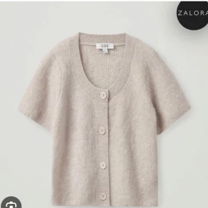 Beige stickad kofta från COS - Superfin beige stickad kofta från COS med korta ärmar och stora knappar framtill. Koftan har en rund halsringning och är gjord i ett mjukt material som känns lyxigt. Perfekt att slänga över en topp för en chill look.