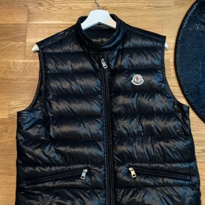 Moncler Gui Väst - Snygg svart dunväst från Moncler med glansig finish och klassisk logga på bröstet. Västen har hög krage, två dragkedjefickor framtill och stängs med dragkedja. Perfekt för lager-på-lager och streetstyle. Jag köpte den här på Plick för runt 4000 men den passar inte mig jätte bra så jag säljer den nu några månader senare. Den är i topp skick och har inga skador eller defekter i huvudtaget och ser som ny ut. Bara att skriva vid frågor bilder eller funderingar.