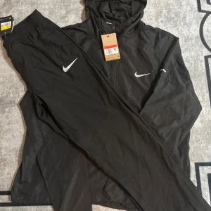 Svart Nike Miller pants  - Svart vindjacka från Nike med huva och dragkedja framtill. Jackan har en stilren look med vit Nike-logga på bröstet och är tillverkad i lätt polyester. Perfekt för löpning eller andra sportiga aktiviteter. Jackan har långa ärmar och en sportig passform.