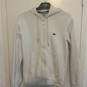 Vit hoodie från Lacoste med dragkedja - Vit hoodie från Lacoste i klassisk modell med dragkedja framtill och huva med snörning. Tröjan har två fickor fram och den ikoniska gröna krokodilloggan på bröstet. Perfekt för chill dagar eller när du vill ha en clean look.
