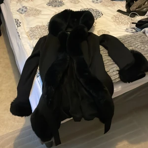 Svart pälsjacka från Garoff med huva - Snygg svart pälsjacka från Garoff med stor fluffig huva och pälsdetaljer vid ärmslut och längs öppningen. Jackan har bälte i midjan och dragkedjor vid fickorna. Perfekt för kalla dagar när du vill vara både varm och stilren.