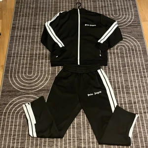 Palm Angels svart tracksuit - Svart tracksuit från Palm Angels med vita ränder längs ärmarna och vit dragkedja framtill. Tröjan har klassisk sportig look, ribbade muddar och logga på bröstet. Perfekt för en streetwear-stil.