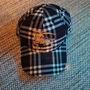 Mörkblå Burberry keps  - Snygg keps från Burberry i klassiskt rutigt mönster i svart, vitt och blått med röda detaljer. Framsidan har en guldfärgad brodyr med Burberry-logga och riddare. Justerbart spänne bak i guldmetall med Burberry-gravyr. Perfekt accessoar för en trendig look. Köpt begagnad och säljer pga att jag ogillar färgen.