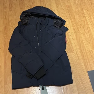 Mörkblå dunjacka från Canada Goose - Säljer en mörkblå dunjacka från Canada Goose med huva och klassisk logga på ärmen. Jackan har långa ärmar, dragkedja och knappar framtill samt ribbade muddar vid ärmslut. Perfekt för kalla dagar och riktigt snygg streetstil.