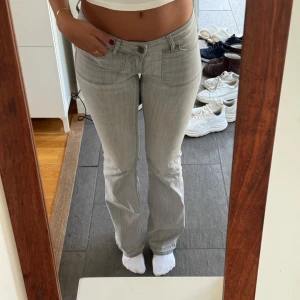 Lågmidjade ljusgrå bootcut jeans med fickdetaljer - Snygga lågmidjade ljusgrå jeans med bootcut passform och låg midja. Jeansen har framfickor med markerade sömmar och bakfickor med lock och knapp. Tillverkade i mjukt denimtyg som sitter skönt.