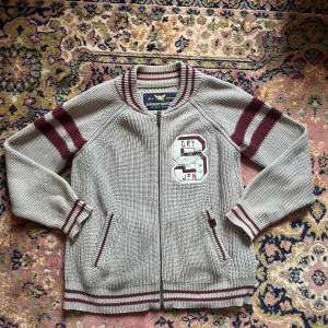 Beige stickad cardigan från Superdry - Säljer en beige stickad kofta från Superdry med vinröda ränder på ärmar och krage. Koftan har dragkedja, två fickor och ett stort collegeinspirerat broderi på bröstet. Perfekt för dig som gillar sportig och retro stil.