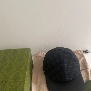 Svart Gucci keps med GG-mönster - Exklusiv svart keps från Gucci med ikoniskt GG-mönster över hela kepsen. Justerbar rem baktill i svart läder med präglad Gucci-logga. Kepsen har en böjd skärm och är tillverkad i bomull med detaljer i läder. Perfekt för dig som vill ha en lyxig streetstyle-accessoar.