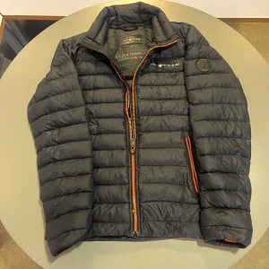 Marinblå dunjacka från Sail Racing - Säljer en svart dunjacka från Sail Racing med orange detaljer på dragkedjorna. Jackan har quiltad design, hög krage och två sidofickor med dragkedja. Perfekt för dig som gillar sportig och clean stil. Insidan har snygga Sail Racing-prints.