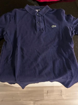 Marinblå pikétröja från Lacoste - Klassisk marinblå pikétröja från Lacoste med den ikoniska broderade krokodilloggan på bröstet. Tröjan har krage, knappar framtill och korta ärmar. Perfekt för en stilren och avslappnad look.