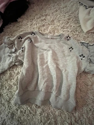 Grå sweatshirt med volangärm Lindex - Grå sweatshirt från Lindex med volangärmar och små broderade blommor i svart och vitt på axlar och ärmar. Tröjan har rund halsringning och ribbade muddar vid ärmslut och nederkant. Perfekt för en avslappnad och trendig look.