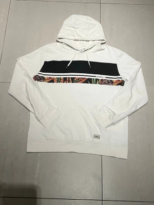 Vit hoodie från O'Neill med tryck - Snygg vit hoodie från O'Neill med svart och färgglad rand över bröstet. Hoodien har huva med dragsko och en stor magficka. Materialet är mjukt och bekvämt, perfekt för en avslappnad stil. O'Neill-loggan syns på bröstet och vid fickan.