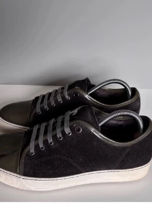Lanvins  - Stilrena svarta sneakers med blankt lack på tån och häl, samt ovandel i mocka. Skorna har grå snörning och vit sula. Perfekta för dig som gillar en clean look med lite edge.mer detaljer om du är intresserad 