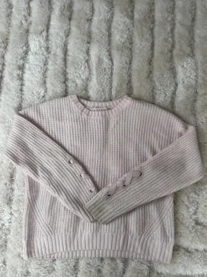 Ljusrosa stickad tröja med snörning - Supersöt ljusrosa stickad tröja med rund hals och ribbade muddar. Ena ärmen har en snygg detalj med snörning i matchande band. Perfekt för dig som gillar en mjuk och trendig look.