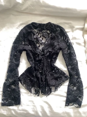 Svart spetsblus med v-ringning - Säljer en svart långärmad blus i genombruten spets med v-ringning och omlottkänsla. Blusen har fina blommiga spetsdetaljer och lätt utsvängda ärmar. Perfekt för dig som vill ha en edgy och elegant look.