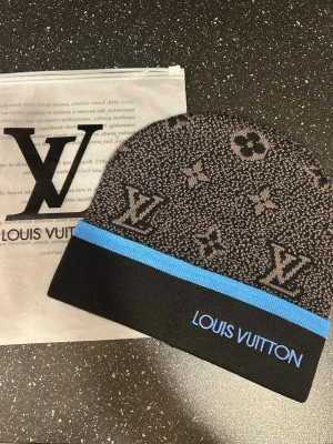 svart mössa med blå rand - Snygg svart mössa med klassiskt monogrammönster i grått och en blå rand längst ner. Märkets logga är broderad i blått på kanten. Perfekt accessoar för dig som vill sticka ut med lyxig streetstyle.