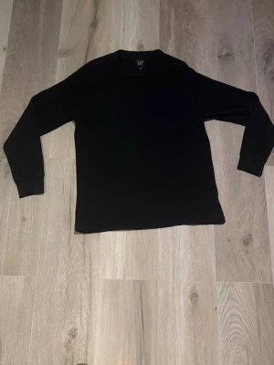 Svart långärmad tröja från GAP - En stilren svart långärmad tröja från GAP i ribbad bomull. Tröjan har rund halsringning och en enkel, klassisk passform som funkar till det mesta. Perfekt basplagg för garderoben.