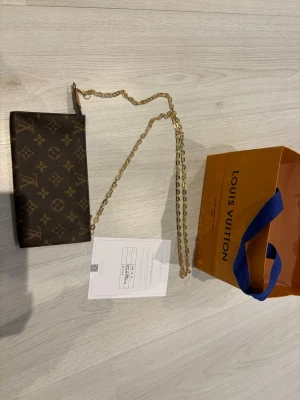 Louis Vuitton brun monogram kuvertväska - Säljer en klassisk brun kuvertväska från Louis Vuitton med ikoniskt monogrammönster och guldfärgad kedja. Väskan är i skinn och har en elegant och stilren design som passar perfekt till din outfit. Kommer med originalpåse och certifikat. Kedjan ej Lv