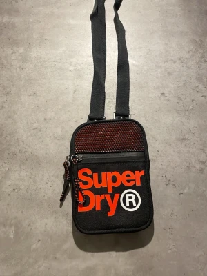 Svart axelväska från Superdry - Snygg svart axelväska från Superdry med röd meshdetalj och stort logotryck i orange framtill. Väskan har justerbar axelrem, dragkedja och praktiskt ytterfack med Superdry-tryck även på baksidan. Perfekt för att bära det viktigaste.