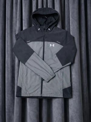 Under Armour jacka - Snygg vindjacka från Under Armour i grått och svart med dragkedja framtill och huva. Jackan har logga på bröstet och justerbara muddar. Perfekt för blåsiga dagar och sportiga outfits.