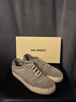 Grå mocka sneakers från Axel Arigato - Snygga grå sneakers från Axel Arigato i mjuk mocka med klassisk låg profil och vita snören. Skorna har en platt sula och minimalistisk design som passar perfekt till streetwear och casual outfits.