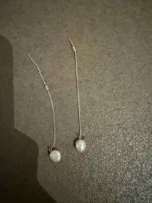 Silvriga örhängen med pärlor - Säljer ett par tunna, silvriga örhängen med hängande vita pärlor. Kedjeformade och minimalistiska, perfekta för dig som gillar stilrena smycken med en touch av elegans. Passar till många olika looks och är superlätta att bära.