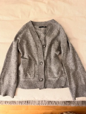 Grå kofta från 157 med fickor - Mysig grå kofta från 157 med knappar framtill och två stora fickor. Koftan är stickad med breda ribbade muddar vid ärmslut och nederkant. Perfekt för lager på lager och enkel att matcha med olika outfits.