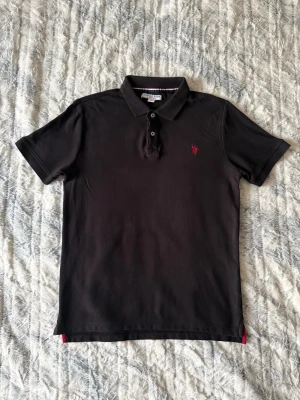 Svart pikétröja från U.S. Polo Assn. - Klassisk svart pikétröja från U.S. Polo Assn. med röd broderad logga på bröstet. Tröjan har korta ärmar, krage och knappar framtill. Tillverkad i bomull för en bekväm känsla. T-shirten är i nyskick och knappt använt. Nypris är 749kr. Strolek M, men passar även S.