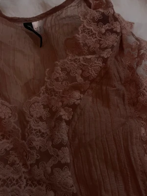 Rosa spetsblus med volangdetaljer - Söt ljusrosa blus från H&M i transparent tyg med spets och volangdetaljer framtill. Blusen är ärmlös och har en v-ringad halsringning med dekorativa spetsmönster längs kanterna. Perfekt för dig som gillar romantisk stil. helt ny aldrig använd. 
