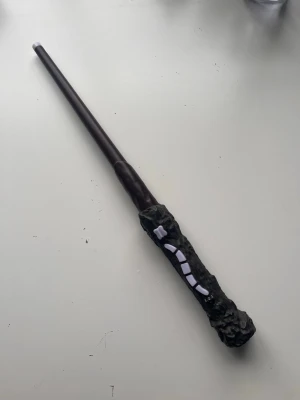 Harry Potter trollstav - Säljer en trollstav inspirerad av Harry Potter-universumet. Staven är mörkbrun med ett detaljerat svart handtag som har vita dekorationer. Perfekt som samlarobjekt.