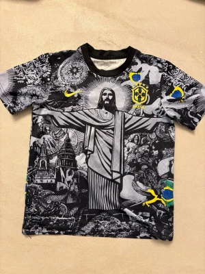 Brasil t-shirt med Kristusmotiv - Unik t-shirt från Nike med svartvitt konstnärligt tryck av Kristusstatyn och brasilianska symboler. T-shirten har korta ärmar, rund hals och detaljer i gult och blått, samt Brasilien-emblem på bröstet. Perfekt för dig som vill sticka ut med en kreativ look. Köpt på nätet för 700 kr