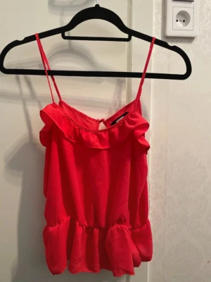 Röd topp - Snygg röd topp med volangdetaljer och smala axelband. Toppen har en söt volangkant upptill och nertill samt en liten öppning med knäppning i ryggen. Perfekt för en trendig och färgstark look. Från bikbok i storlek S.