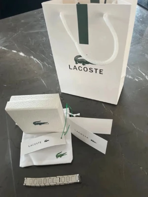 Silverfärgad klocklänk från Lacoste - Snygg och stilren silverfärgad klocklänk från Lacoste. Länken är i metall och har en klassisk design med rektangulära länkar. Perfekt för dig som vill byta ut eller uppgradera din klocka med en exklusiv känsla. Kommer med originalförpackning, påse och tags.