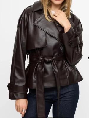 Brun skinnjacka med bälte från Weekday - Snygg mörkbrun skinnjacka från bikbok med klassisk krage, knappar och bälte i midjan. Jackan har långa ärmar och en loose passform som ger en cool och modern look. Perfekt för dig som vill sticka ut med en trendig jacka i skinn. Slutsåld på hemsidan och är i helt nytt skick. Nypris 1199 kr