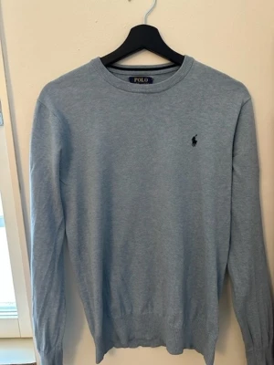 Ljusblå stickad tröja Polo Ralph Lauren - Ljusblå stickad tröja från Polo Ralph Lauren i mjuk pima-bomull. Klassisk rund halsringning och diskret broderad logga på bröstet. Långa ärmar och ribbade muddar vid ärmslut och nederkant. Perfekt för en stilren look.