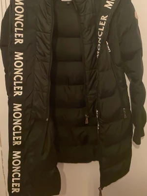 Svart lång dunjacka från Moncler - Snygg svart lång dunjacka från Moncler med stora logotexter längs dragkedjan. Jackan har quiltad design, dragkedja framtill och sidofickor. Perfekt för kalla dagar och ger en riktigt trendig look dock har texten från bild 3 lossnat lite men enkelt att fixa genom att sy